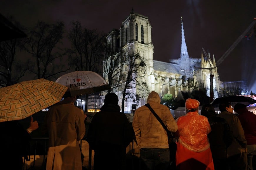 People protect themselves from the rain under umbrellas as they watch on a screen the reopening service ceremony of the Notre-Dame de Paris Cathedral, five-and-a-half years after a fire ravaged the Gothic masterpiece, as part ceremonies to mark the Cathedral's reopening after its restoration, in Paris, France, December 7, 2024.  REUTERS/Kevin Coombs