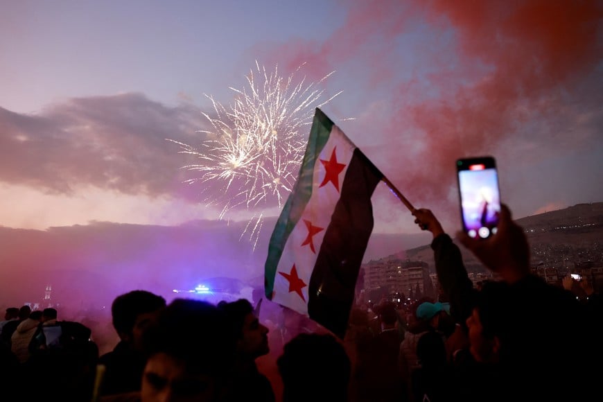 A person waves a flag adopted by the new Syrian rulers, as fireworks explode, while people gather during a celebration called by Hayat Tahrir al-Sham (HTS) near the Umayyad Mosque, after the ousting of Syria's Bashar al-Assad, in Damascus, Syria, December 20, 2024. REUTERS/Ammar Awad