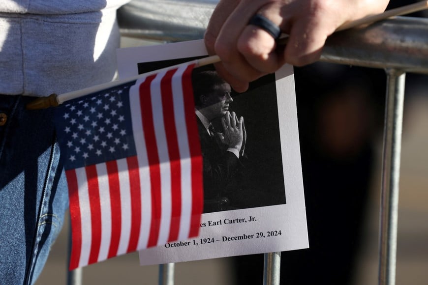 A person holds a U.S. flag and an image of former U.S. President Jimmy Carter while waiting for the motorcade carrying his casket to pass on its way to his private funeral service, in Plains, Georgia, U.S. January 9, 2025.  REUTERS/Sam Wolfe